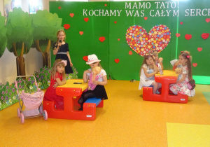 Kilka dziewczynek siedzi na rozstawionych ławeczkach z lalkami, jedna z dziewczynek stoi z boku i trzyma w ręku mikrofon recytuje wiersz.